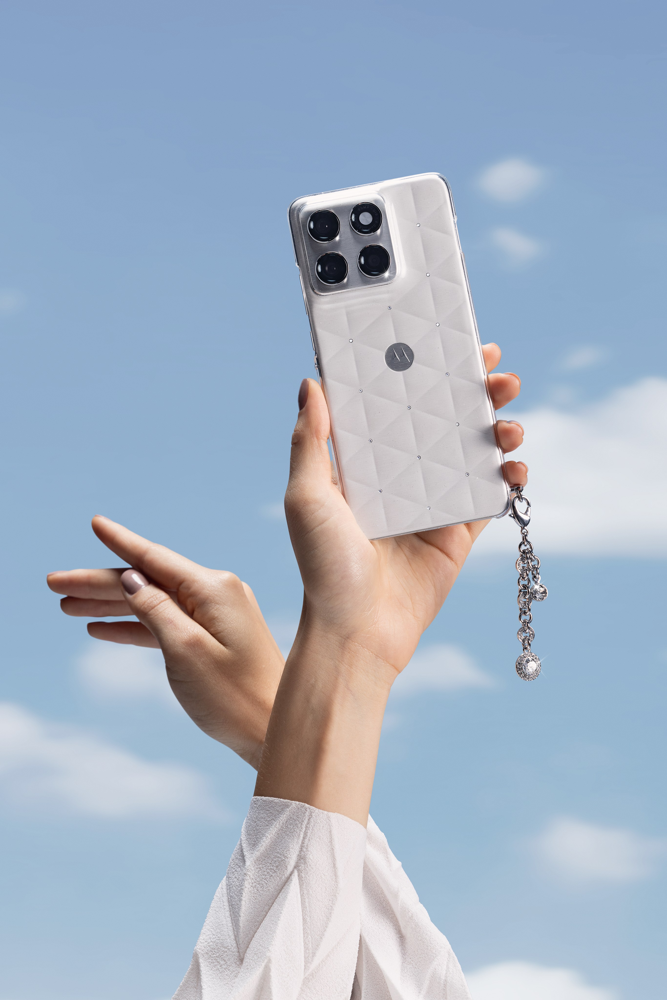Raised hand holds a white quilted-texture motorola phone with a crystal charm, sky behind