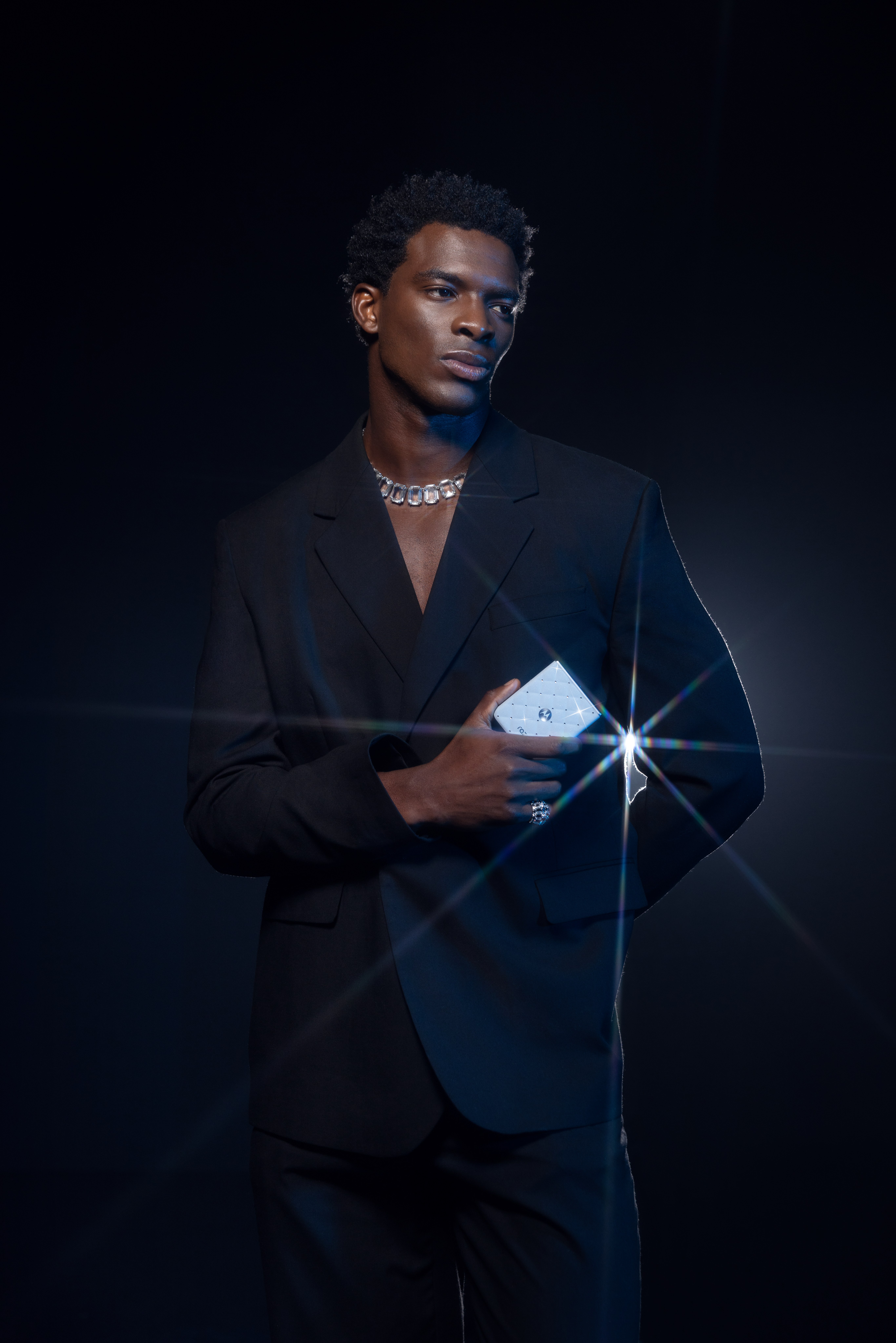 Man in a black suit and crystal necklace holds a sparkling phone to his chest against a dark backdrop