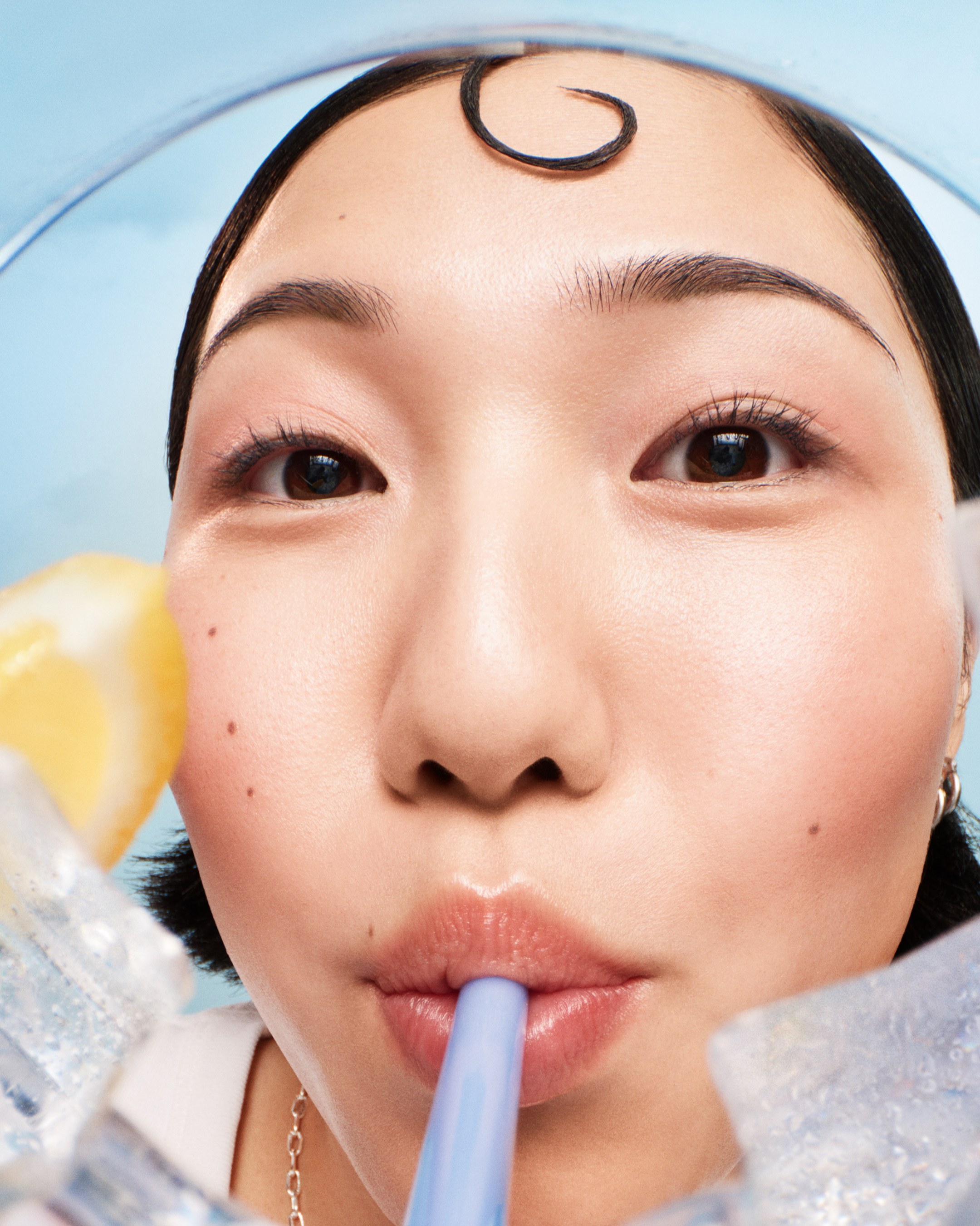Close-up of a woman with a curl of hair on her forehead sipping through a blue straw, seen from inside a glass with ice and lemon