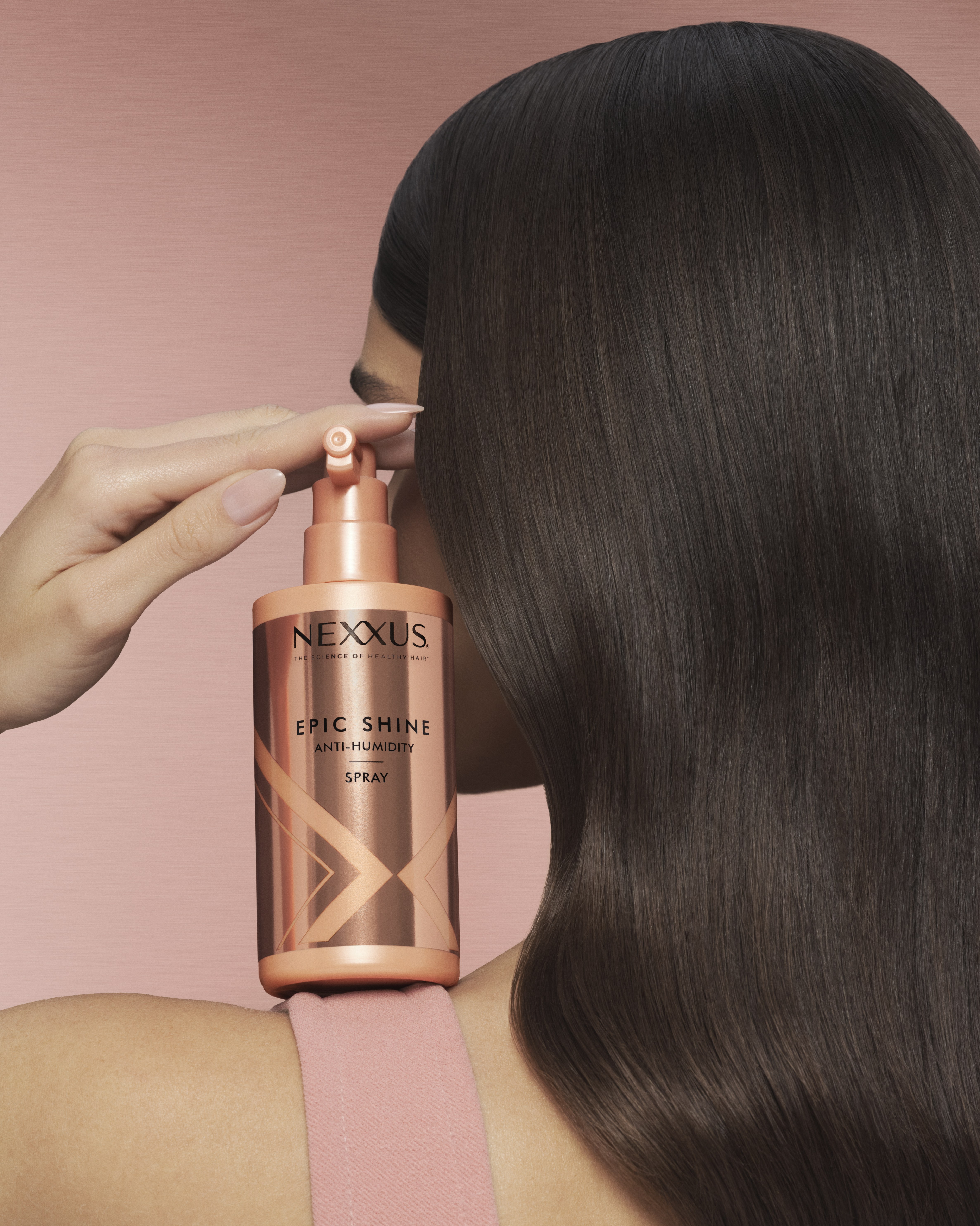 Product shot of a Nexxus Epic Shine anti-humidity spray balanced on a shoulder beside long glossy brown hair, pink backdrop