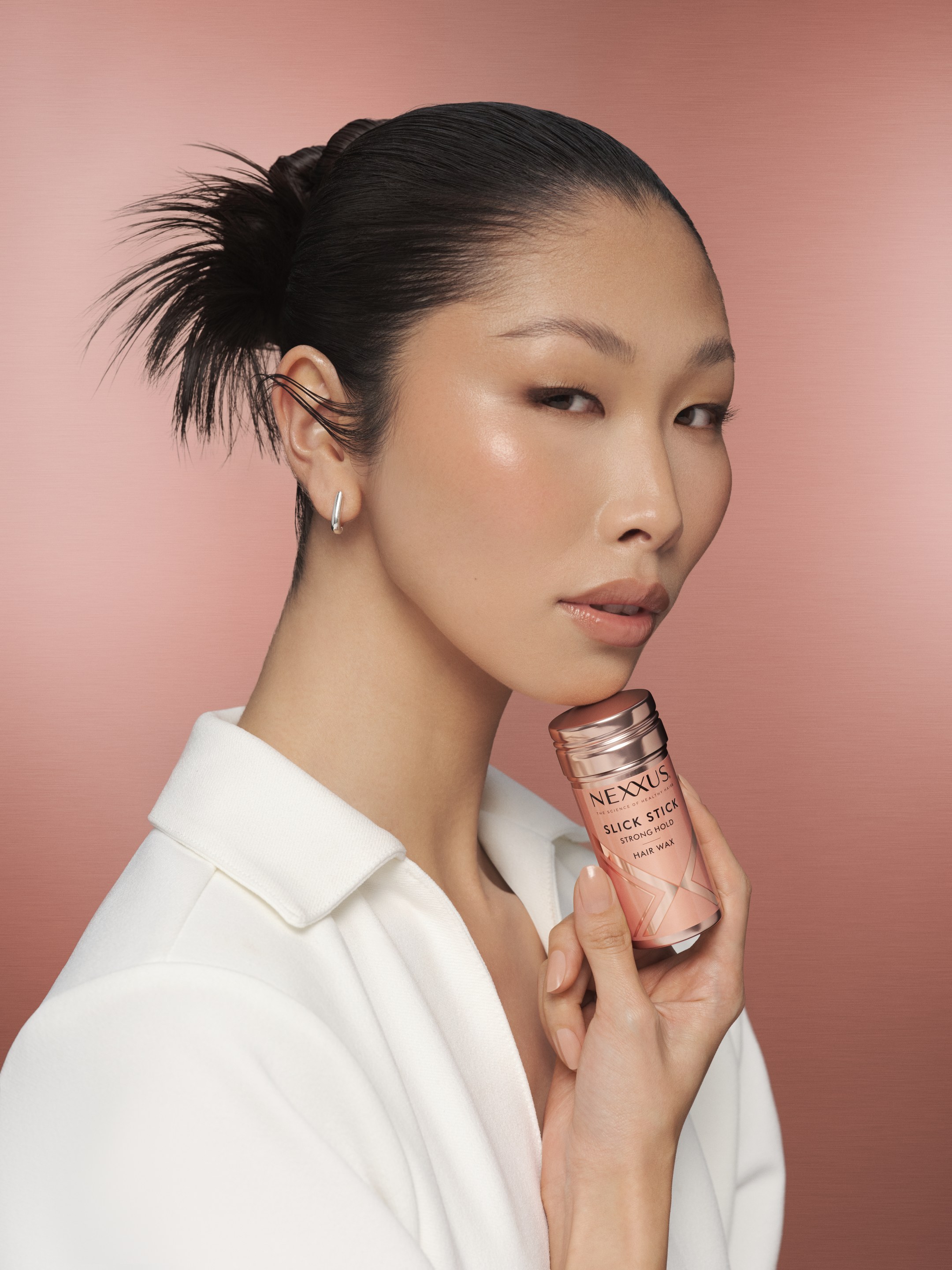 Model with a sleek bun holds a small pink Nexxus bottle to her cheek against a pink backdrop