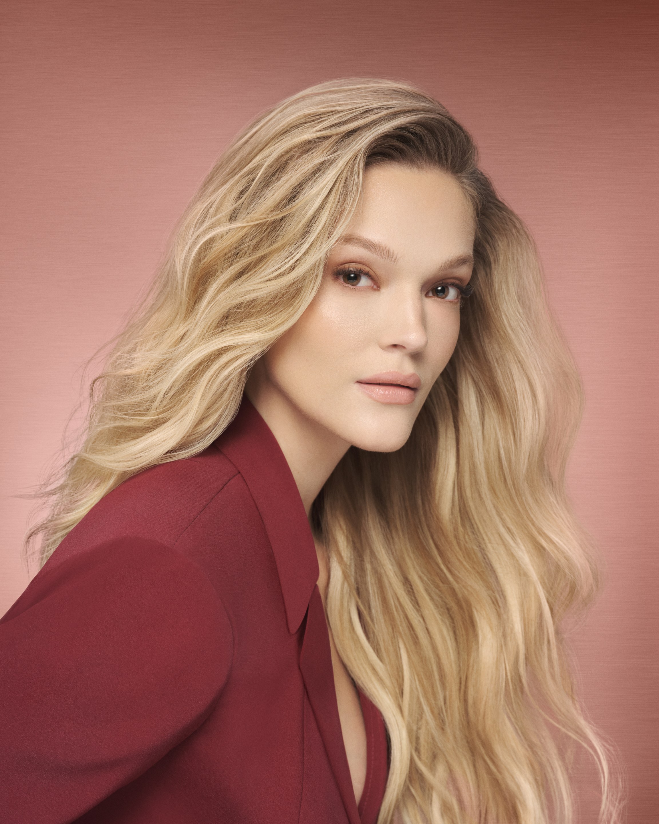 Blonde model in a burgundy blazer looks to camera, long waves over one shoulder, pink backdrop
