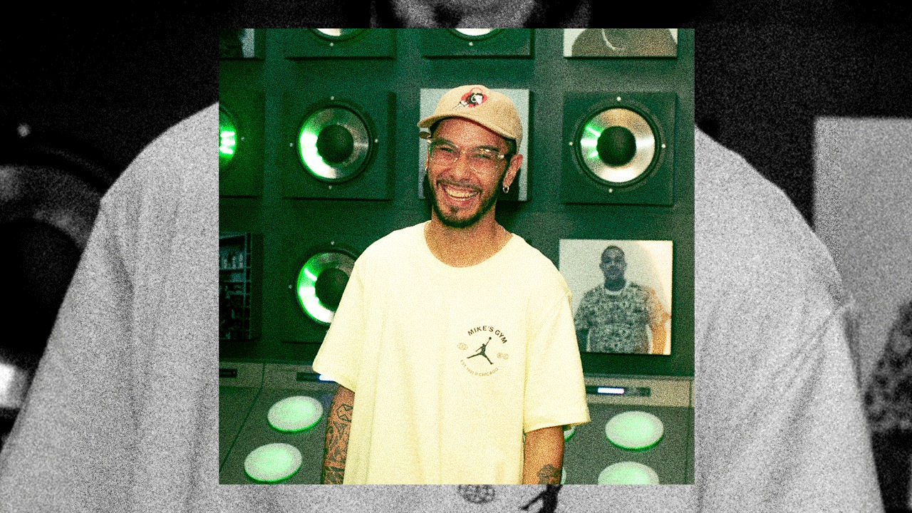 A smiling man in a cap in front of a wall of glowing green speakers and portrait photographs