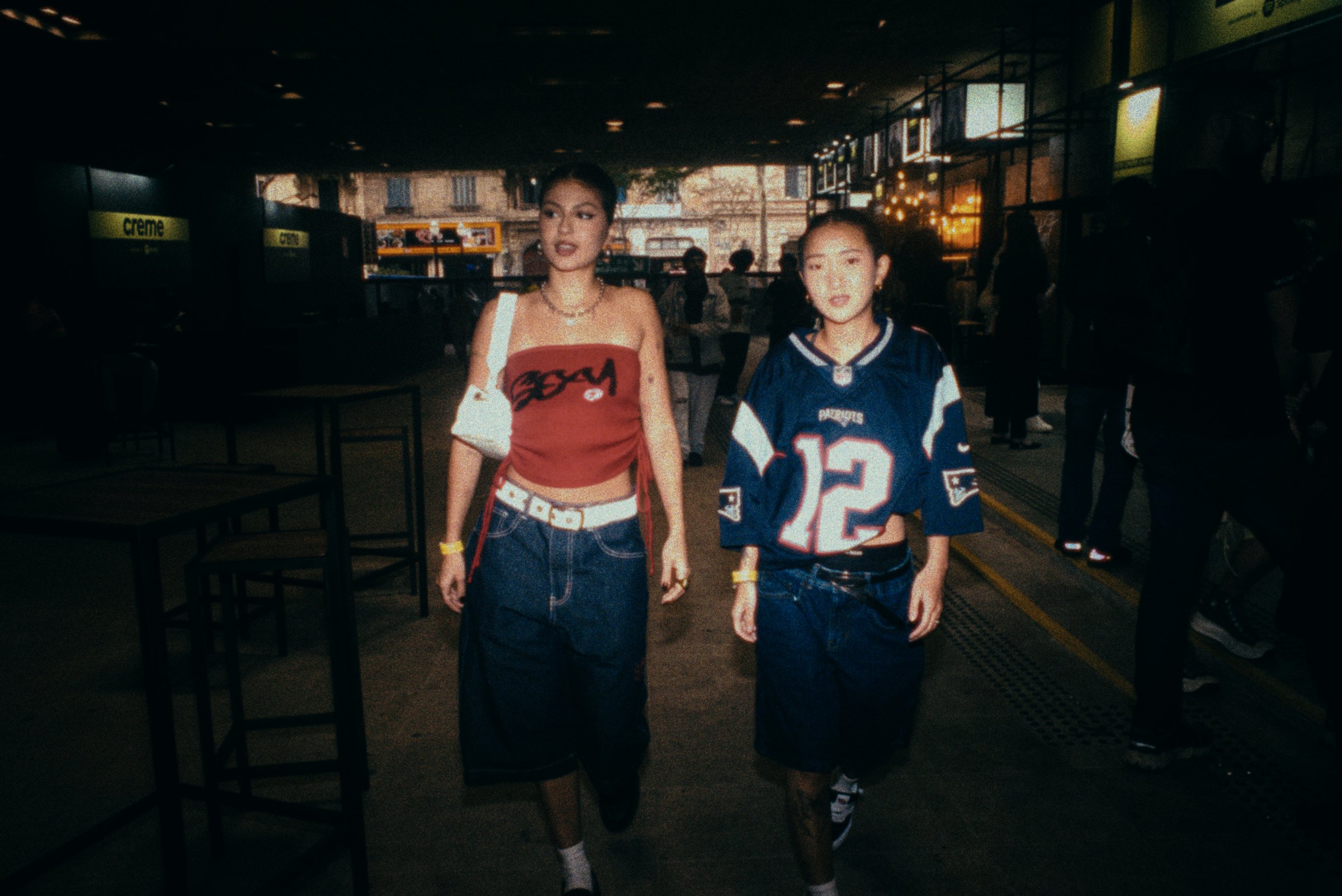 Two young women walk side by side at night, one in a red corset top, one in a football jersey