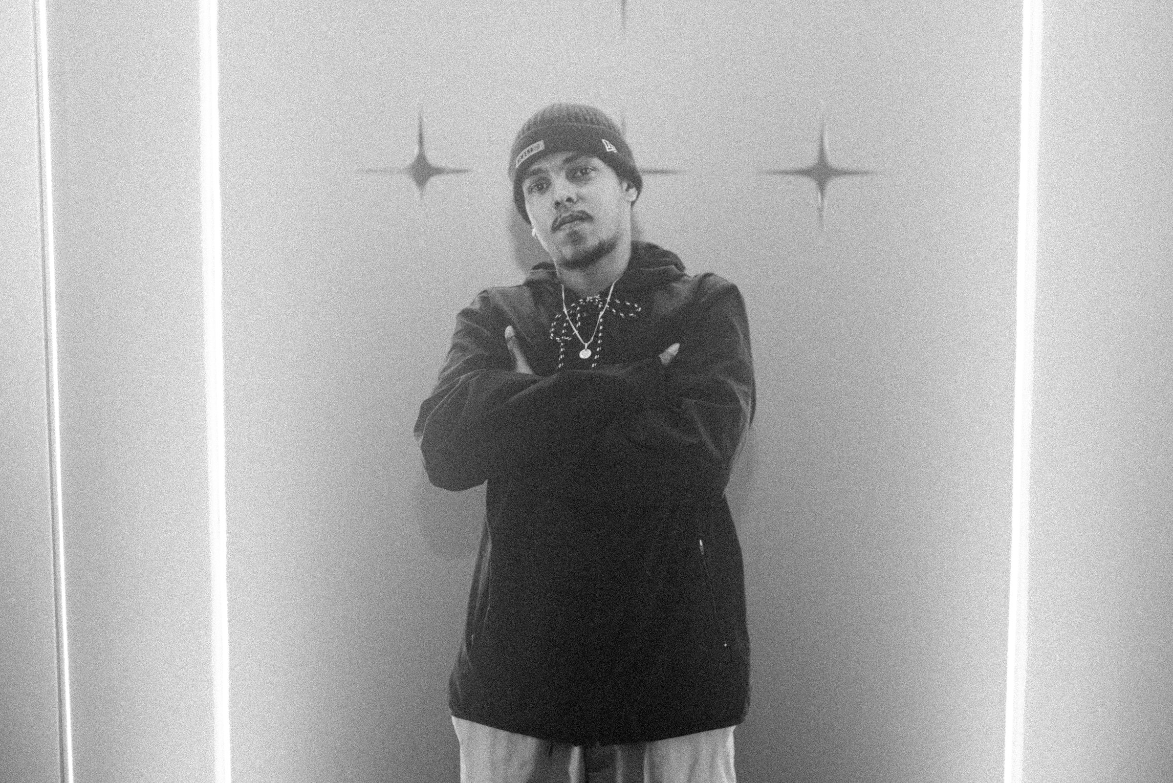 Black and white: a man in a cap and dark jacket stands with folded arms in a white room