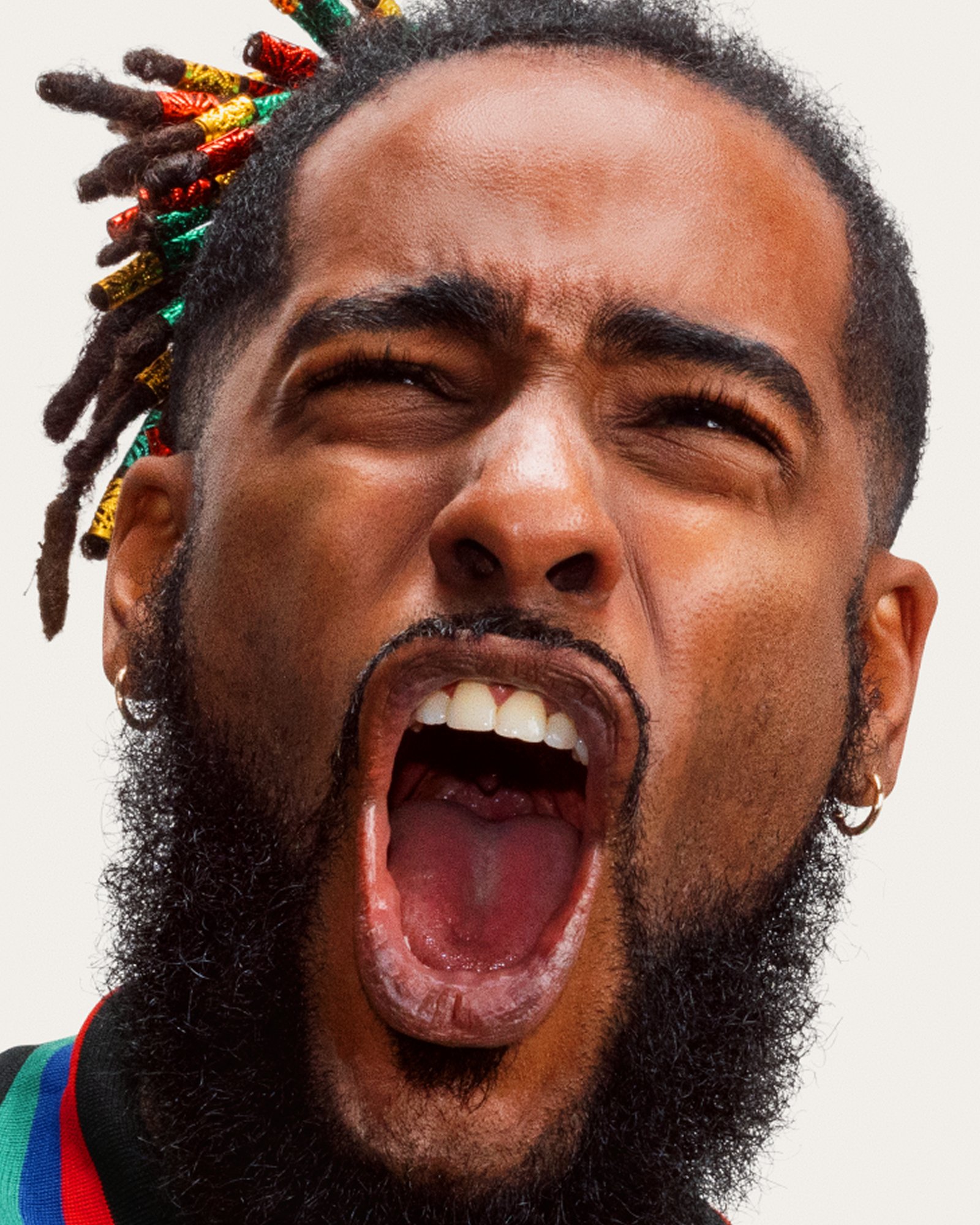 Close-up of a bearded man with colorful wrapped locs shouting with his mouth wide open, on a white backdrop