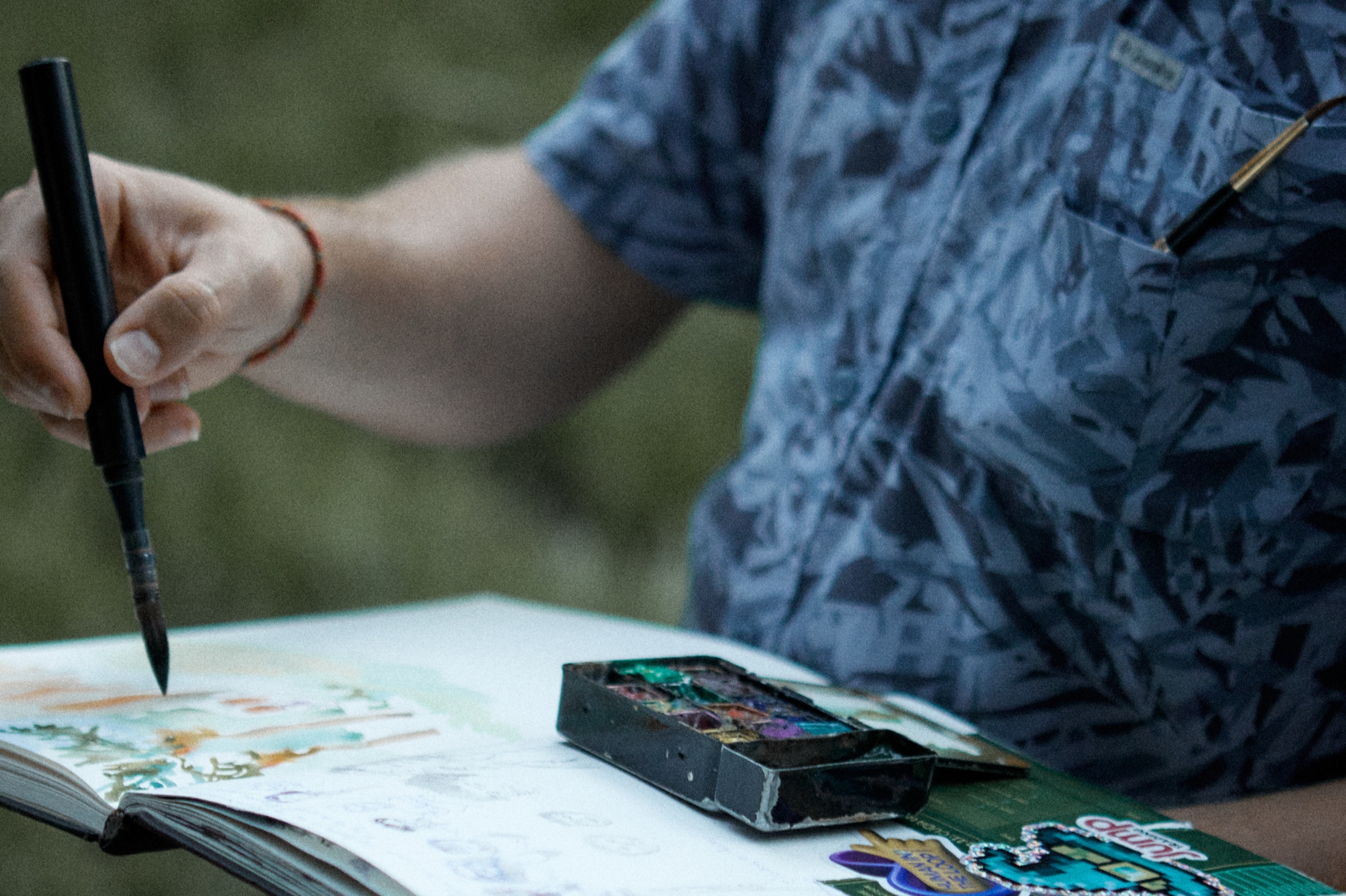 Close-up of a hand painting watercolors in a sketchbook beside a travel paint tin, outdoors