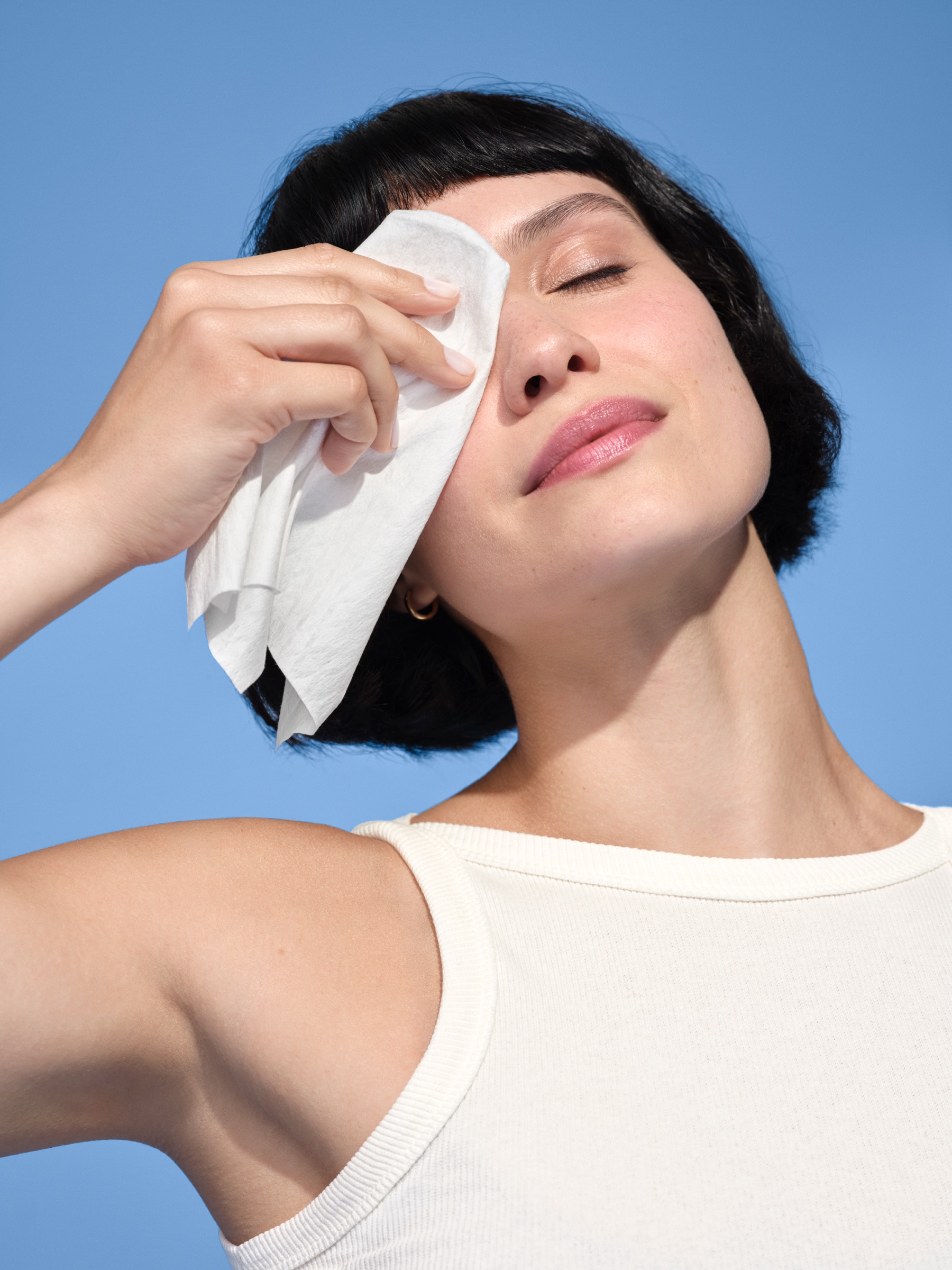 Woman with a black bob tilts her head back and dabs her eye with a white disposable face towel, smiling against a blue sky backdrop