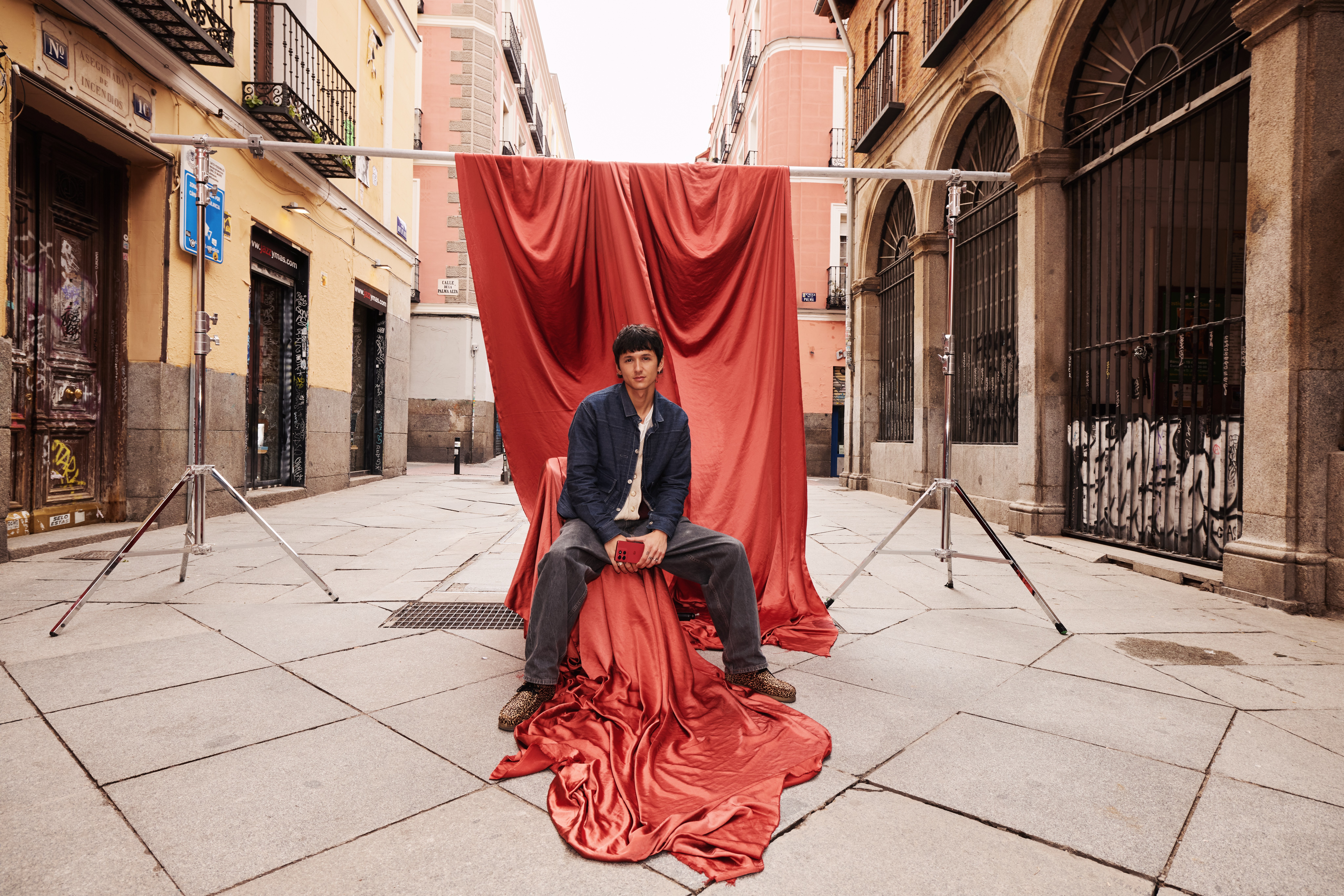 Young man sits on draped red satin in the middle of a stone street holding a red phone, light stands on each side