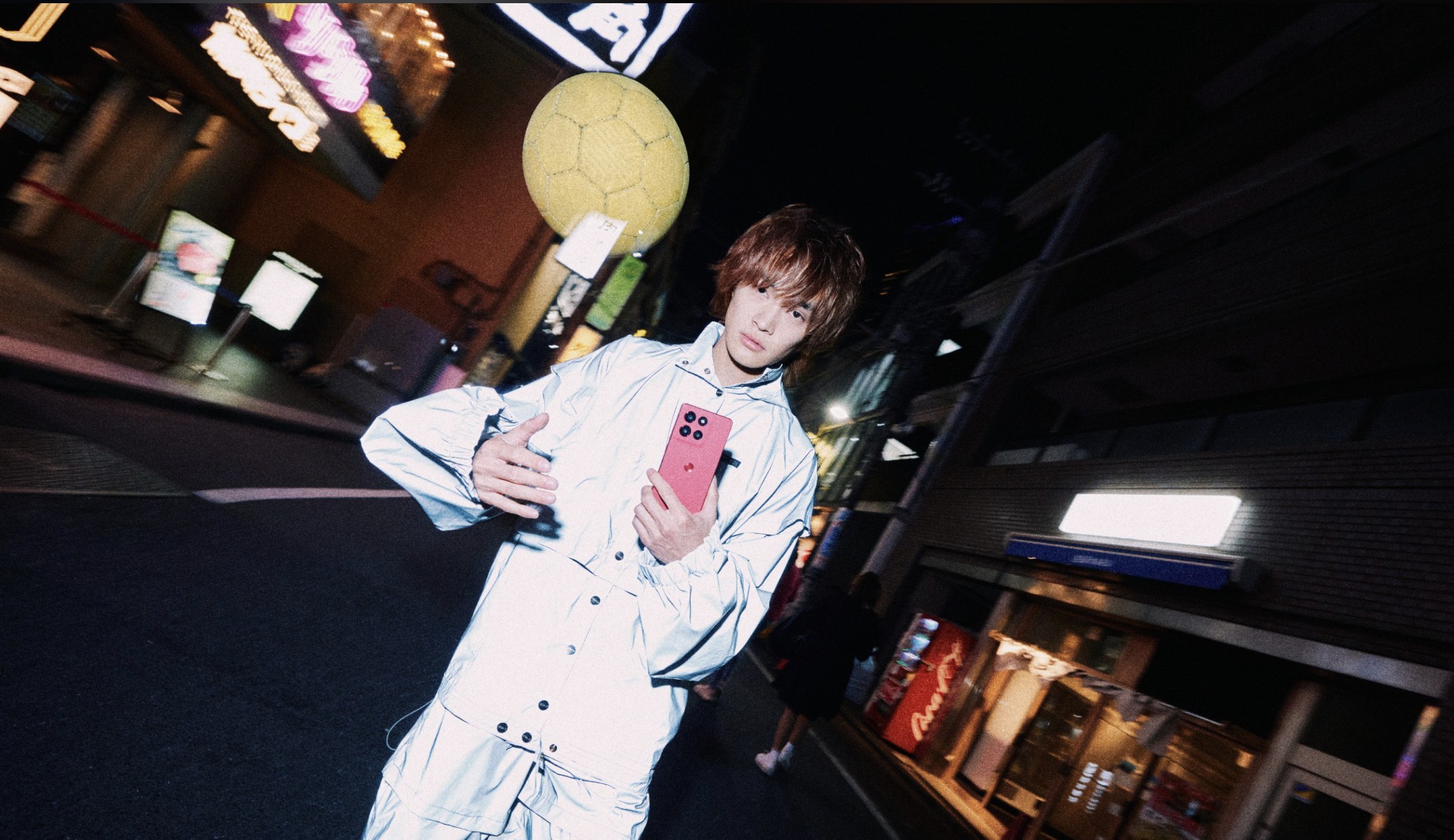 Person in a reflective silver jacket holds a pink phone on a night street under a football-shaped lamp