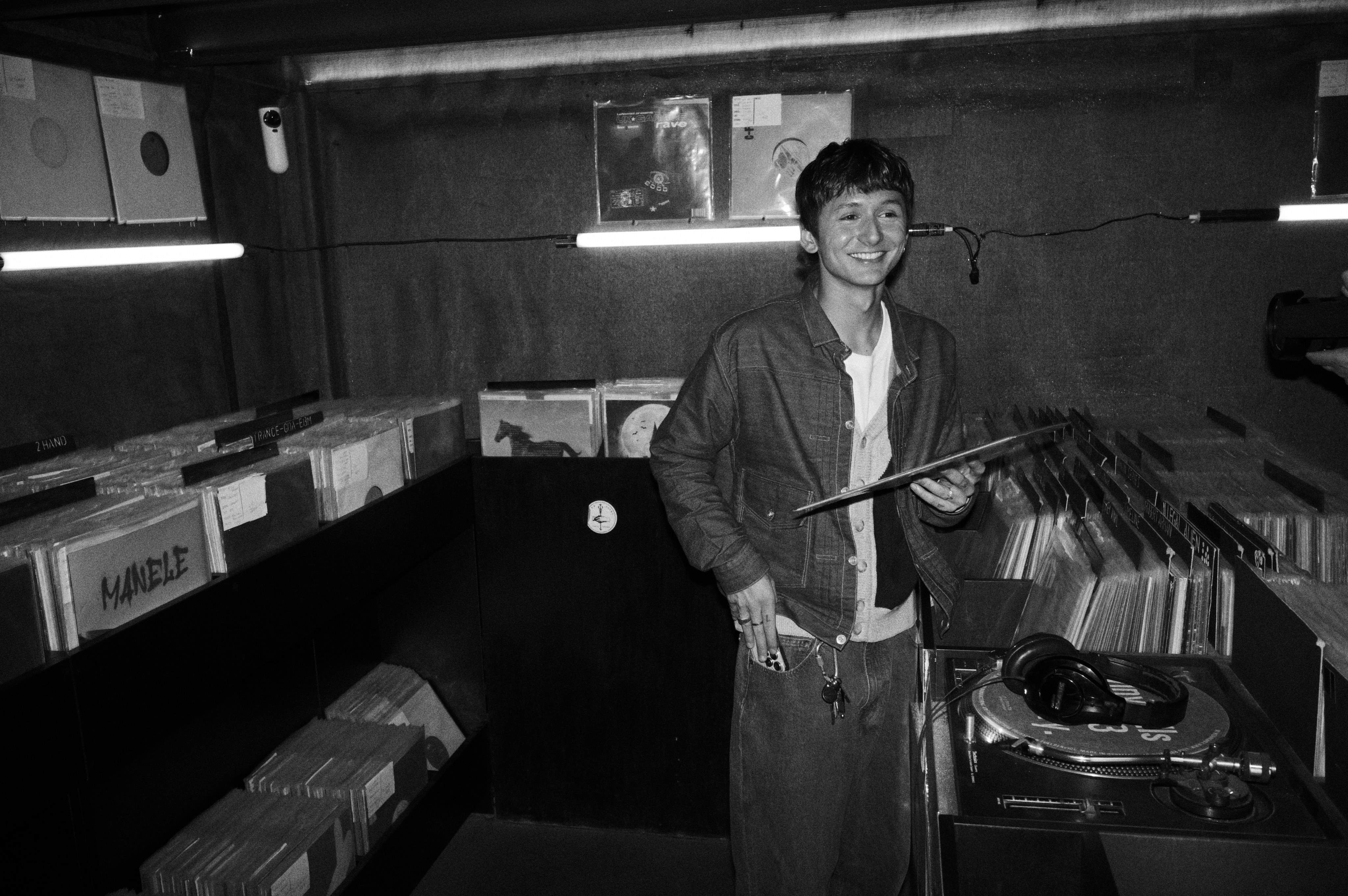 Black and white photo of a smiling young man holding a record in a vinyl shop beside a turntable
