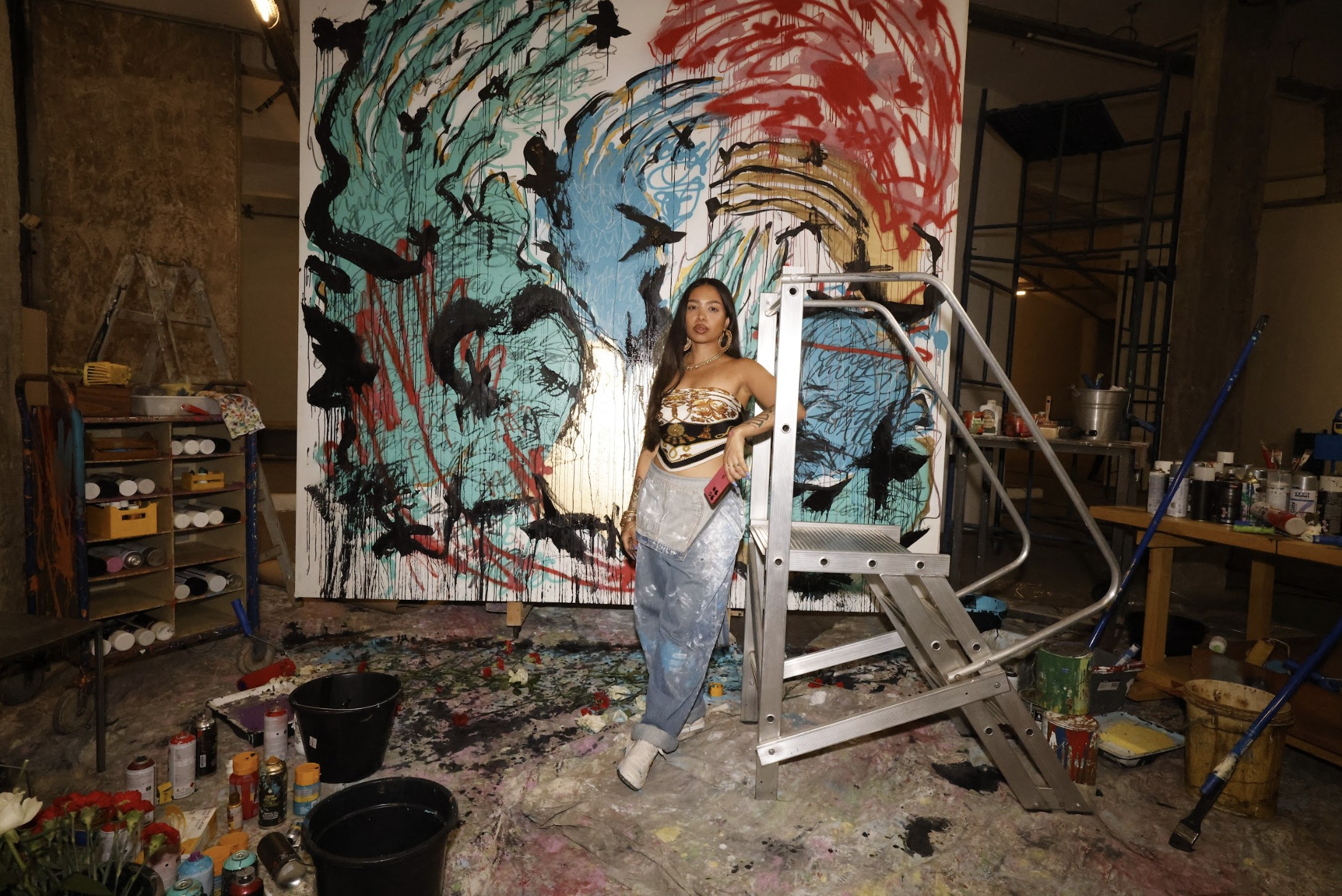 Woman leans on a metal step ladder in a paint-splattered studio in front of a large abstract mural