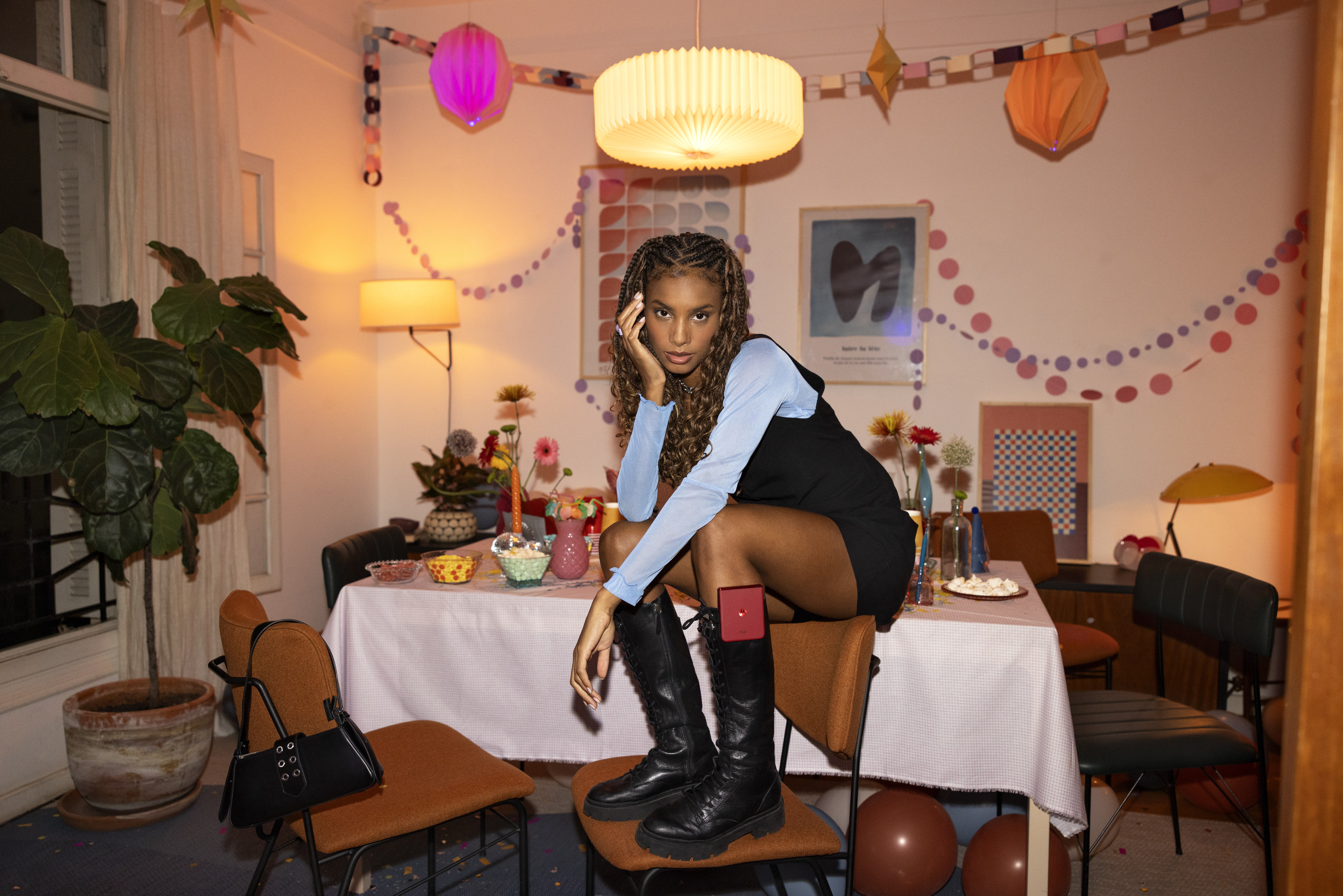 Woman in black boots crouches on a chair at a party table, a pink flip phone tucked in her boot